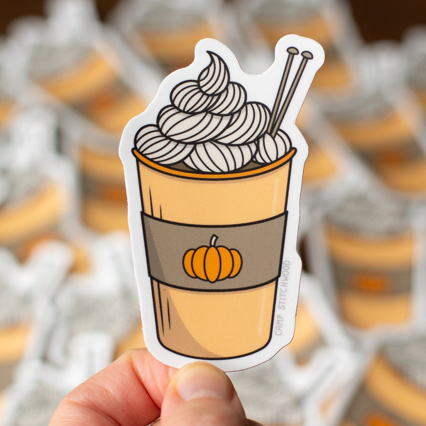 Sticker of a pumpkin spice latte with whipped cream held in front of a blurred background of similar stickers.