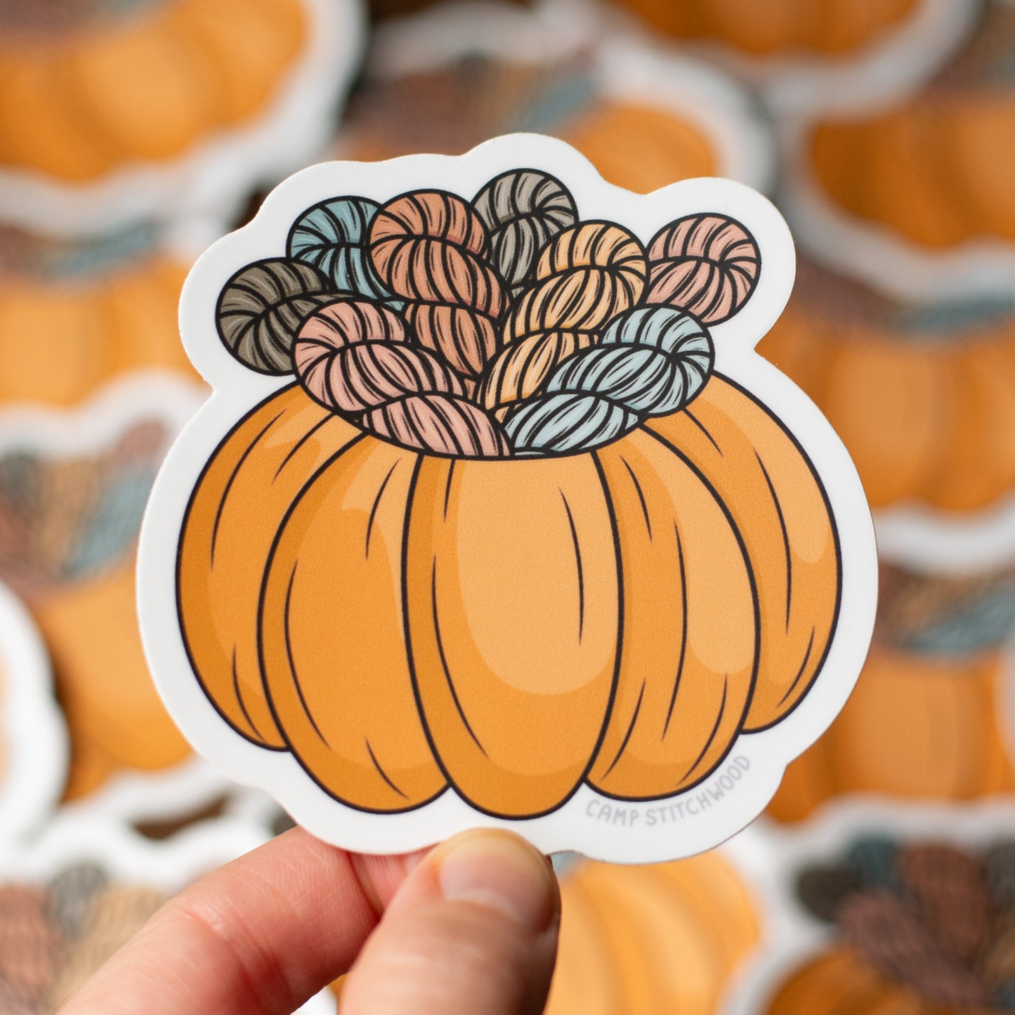 Sticker of a pumpkin with yarn on top held by a hand, with more stickers in the background.