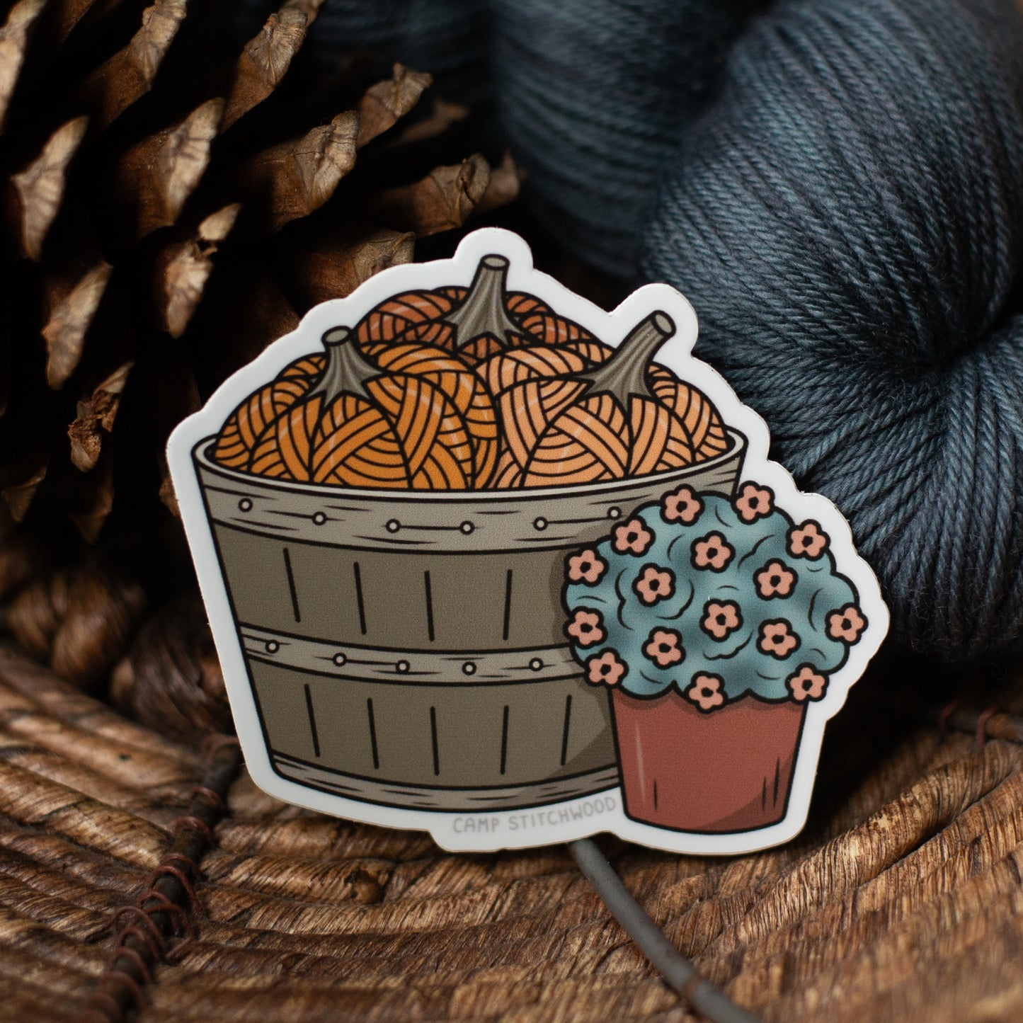 Sticker of a basket with yarn pumpkins and a flower pot on a wooden surface with yarn and pine cones.