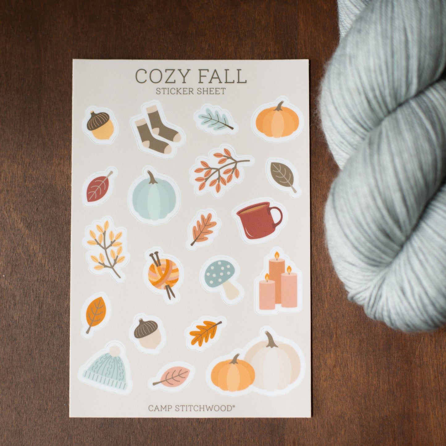 Sticker sheet with autumn-themed designs on a wooden surface next to a skein of yarn.