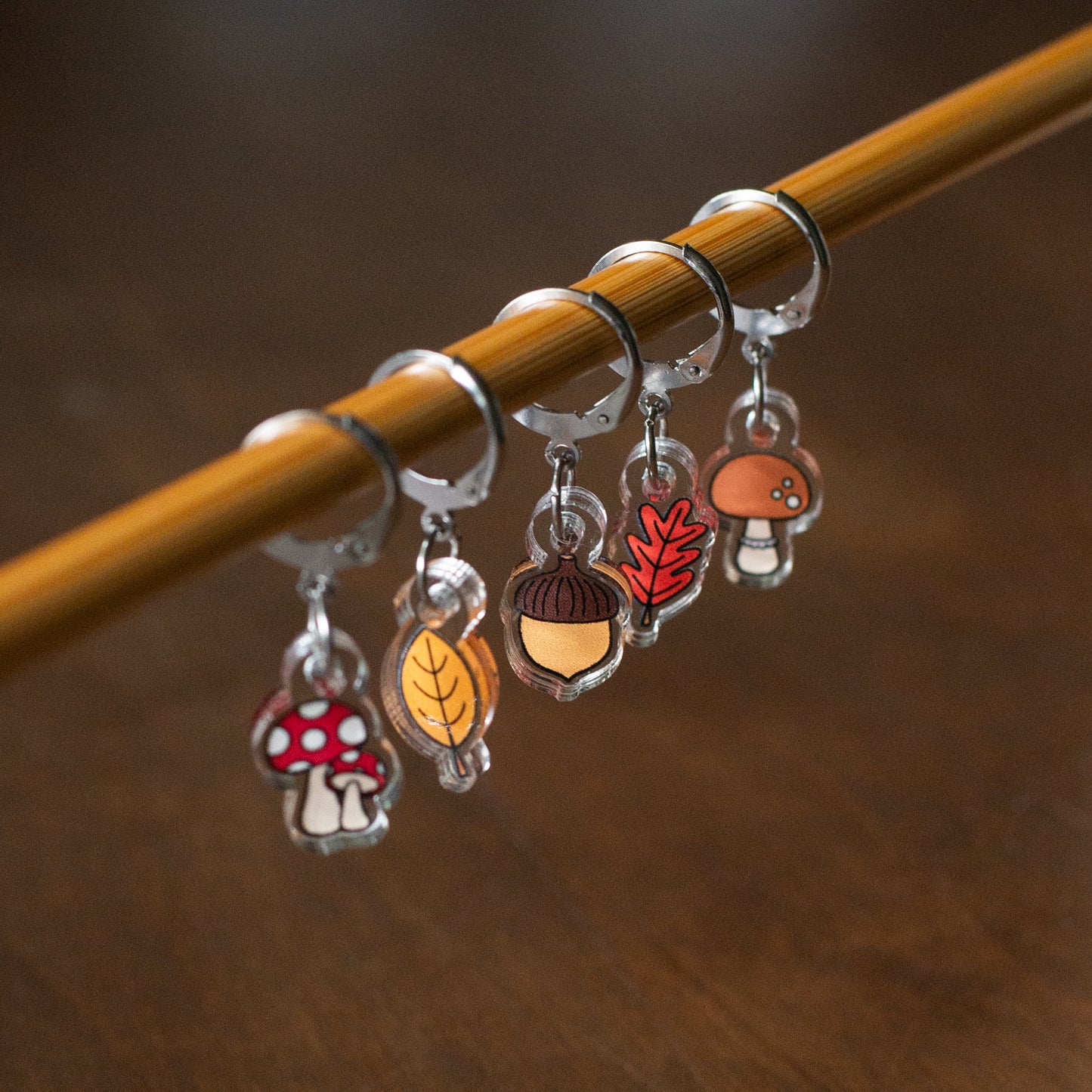 Set of colorful mushroom and leaf stitch markers on a knitting needle against a brown background