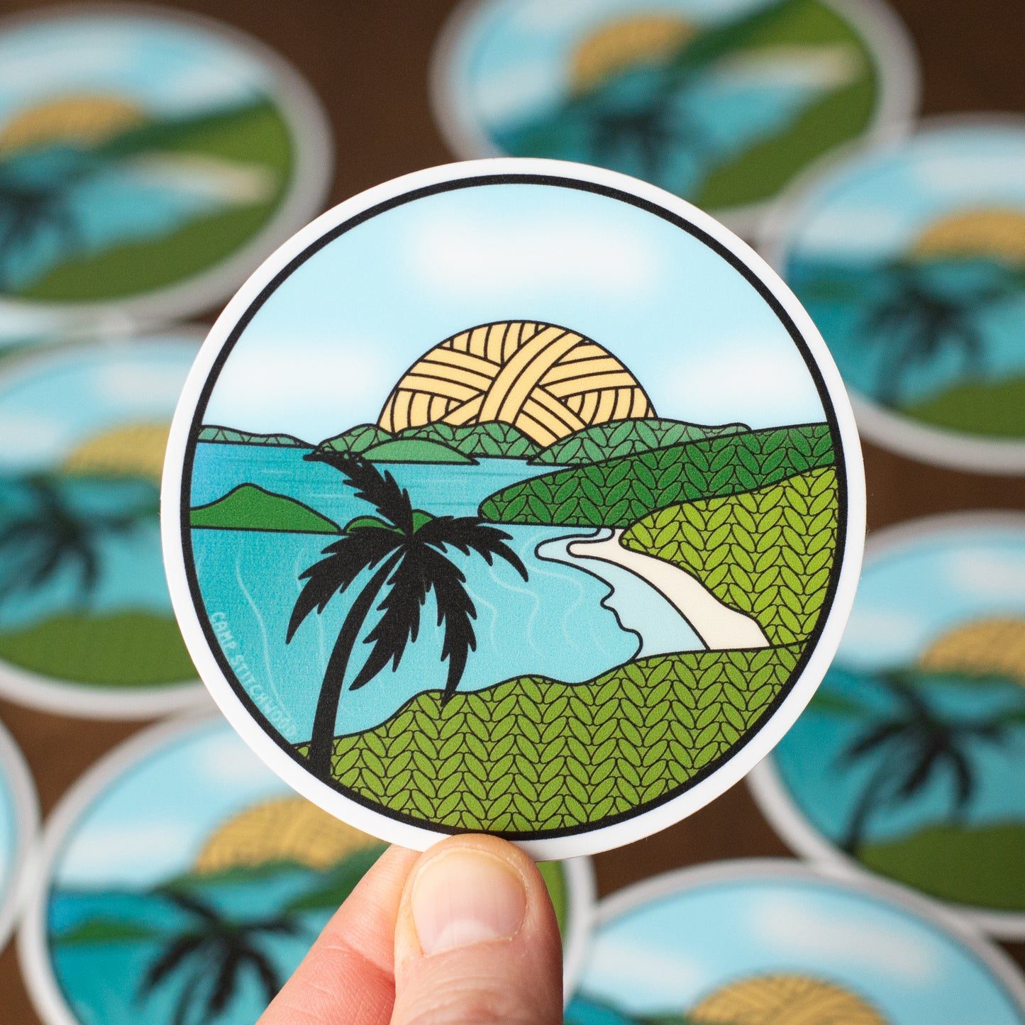 Circular sticker with a tropical design held by a hand, surrounded by more stickers on a brown background.
