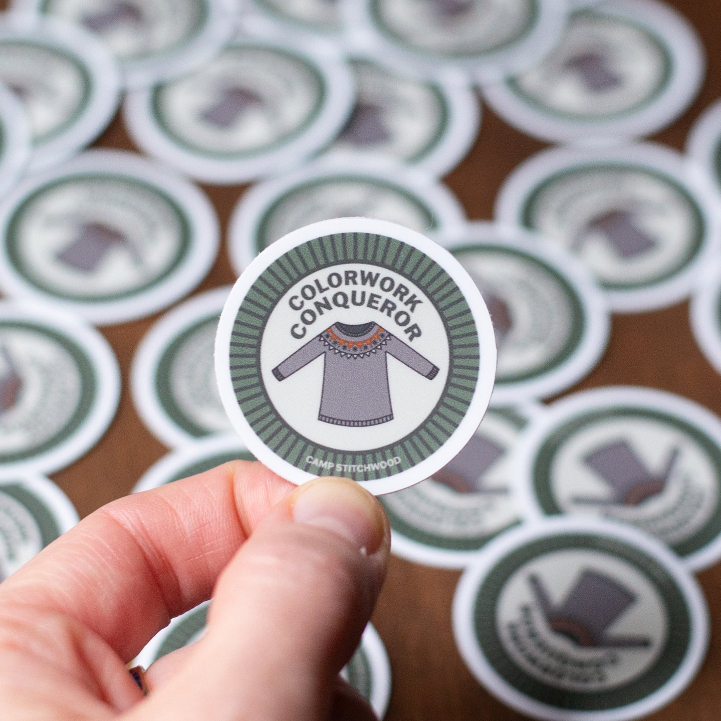 Sticker with 'Colorwork Conqueror' design held by a hand, surrounded by more stickers.