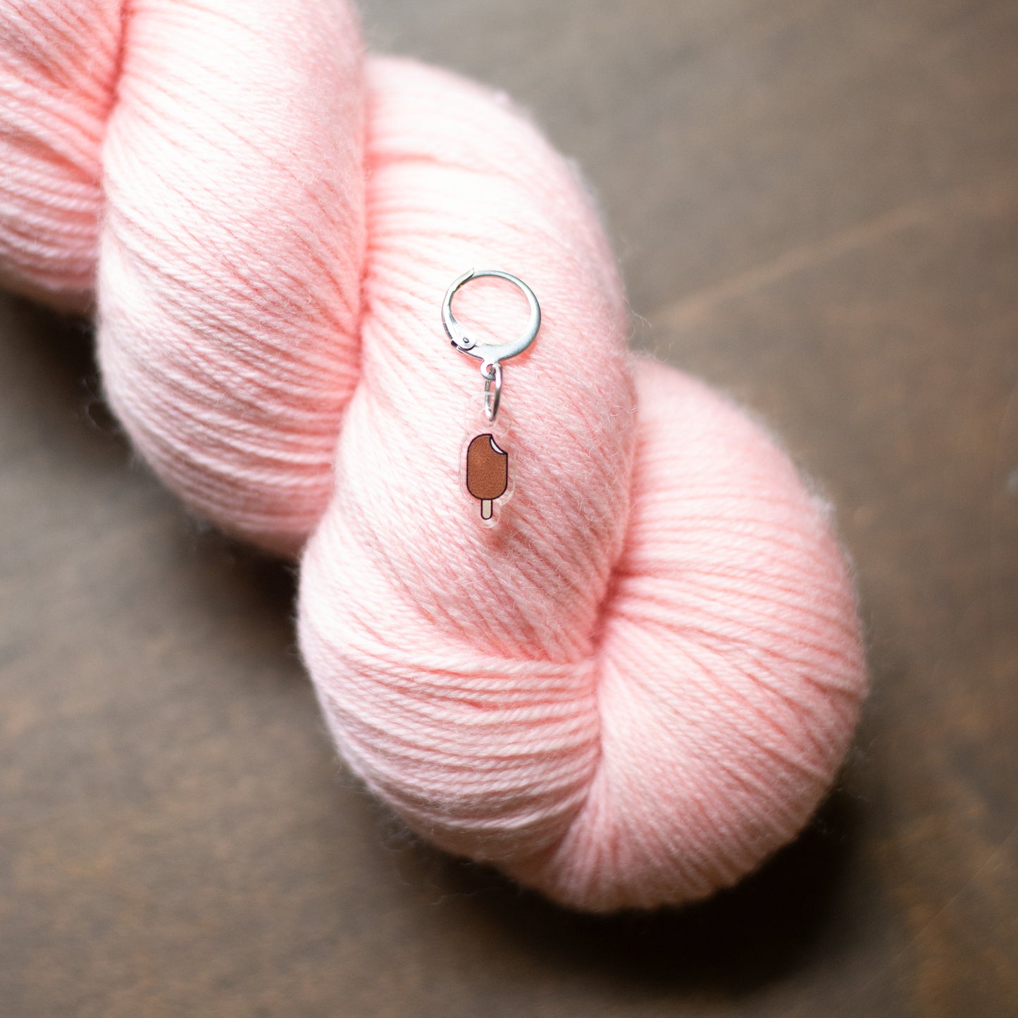 A close-up of a soft pink skein of yarn on a dark brown wooden surface, with a small ice pop stitch marker.