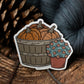 Sticker of a basket with yarn pumpkins and a flower pot on a wooden surface with yarn and pine cones.