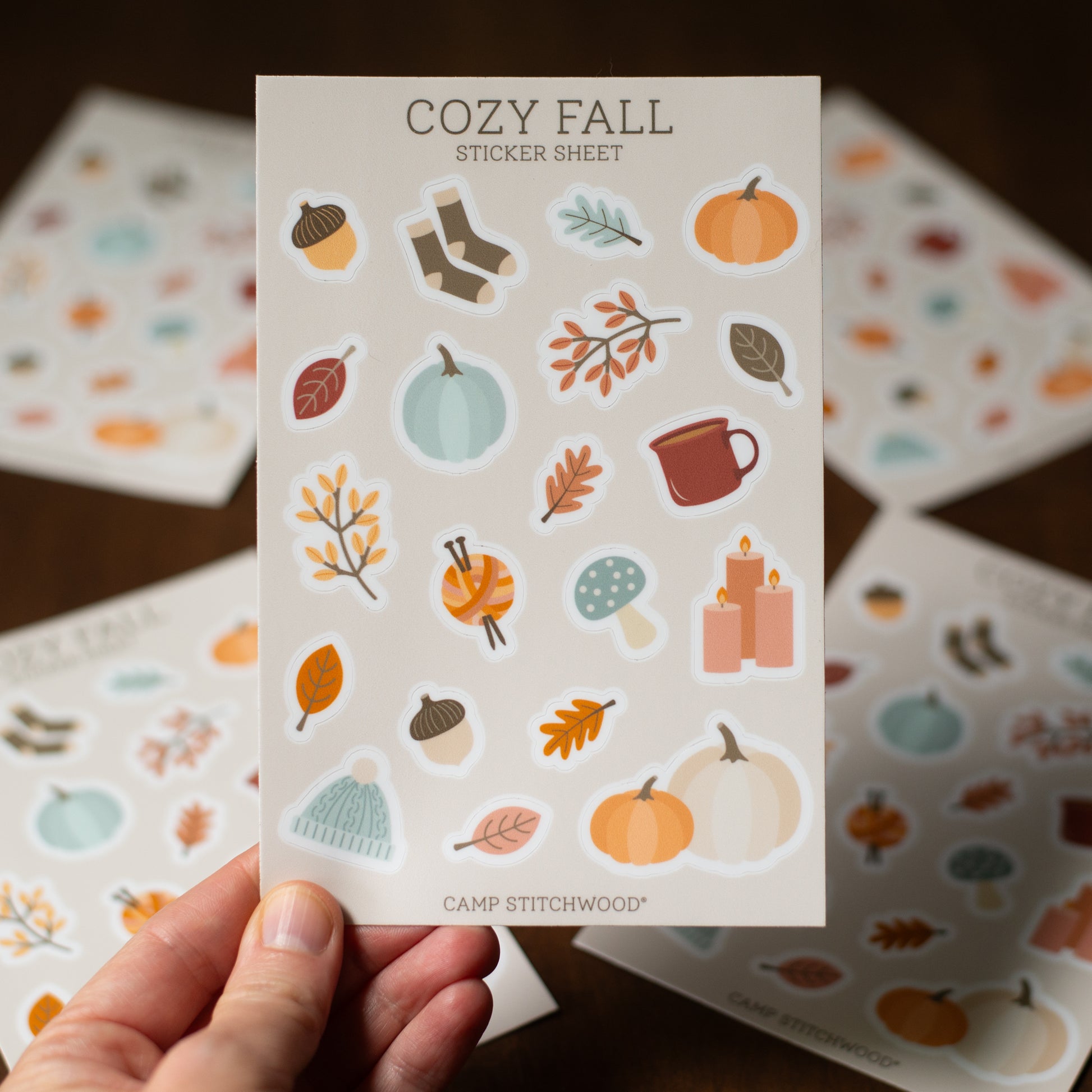 Sticker sheet with fall-themed designs held by a hand, with more sheets in the background.