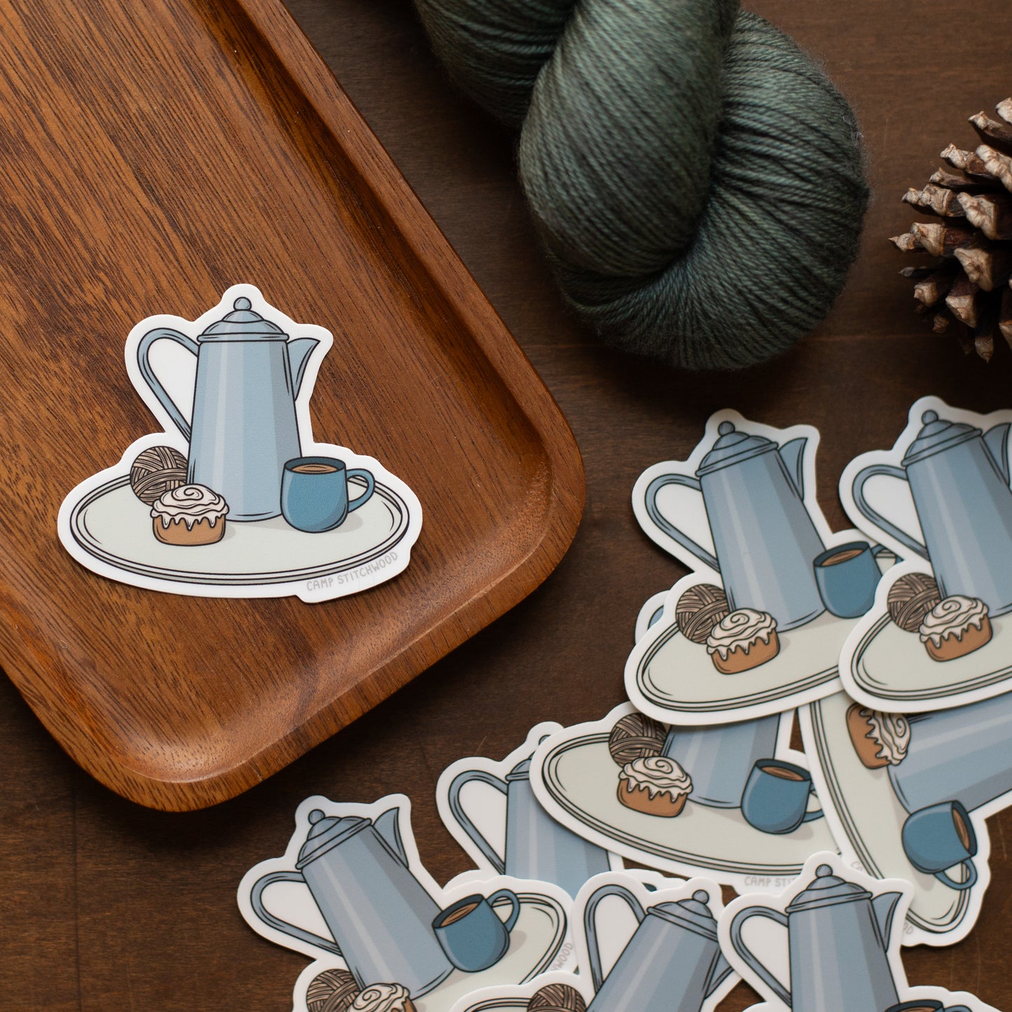 Sticker of a teapot and cup on a wooden tray with yarn and pine cones.