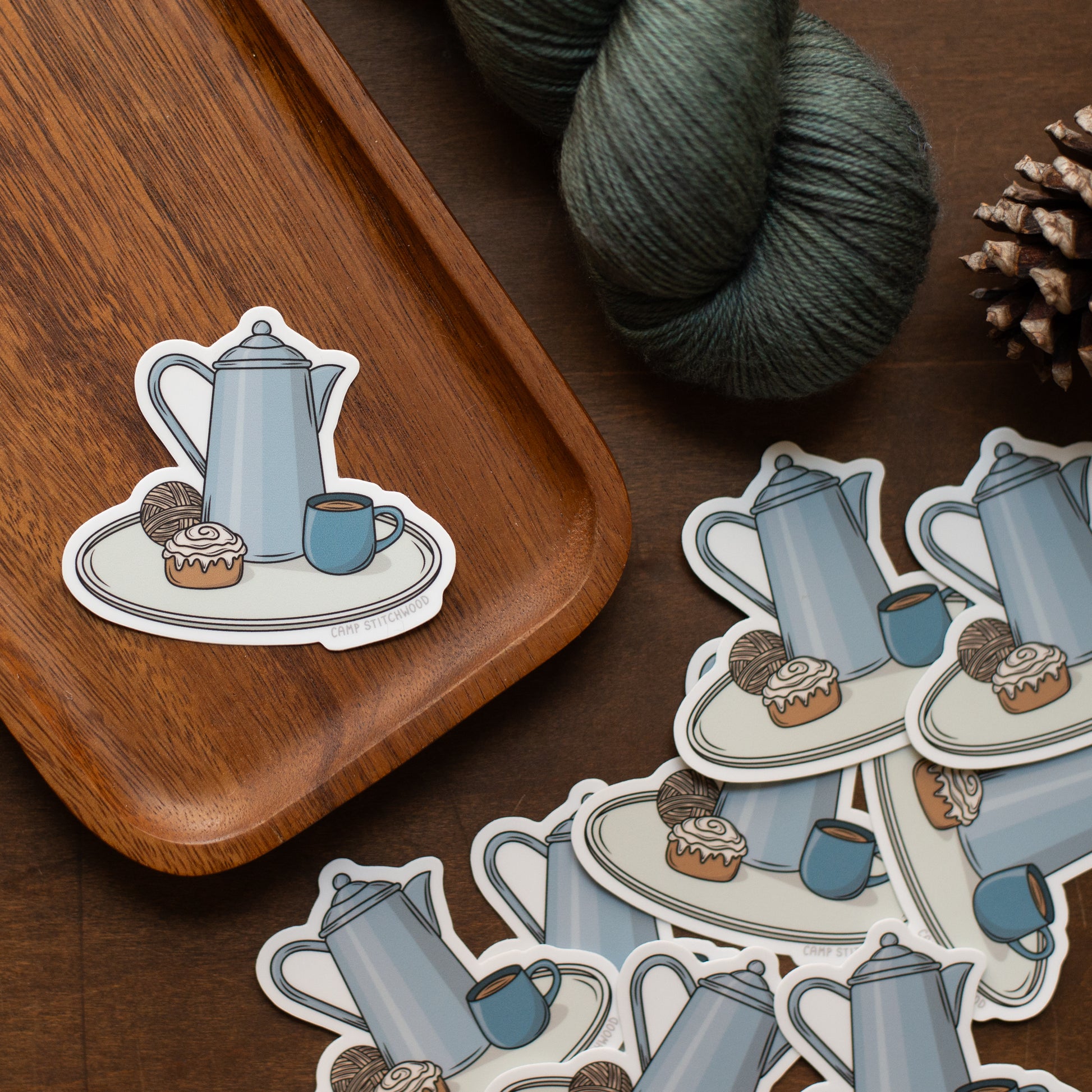 Sticker of a teapot and cup on a wooden tray with yarn and pine cones.
