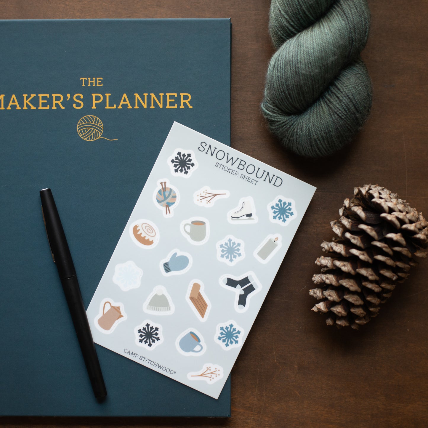 Maker's Planner with a sheet of 'Snowbound' stickers, pen, and yarn on a wooden surface