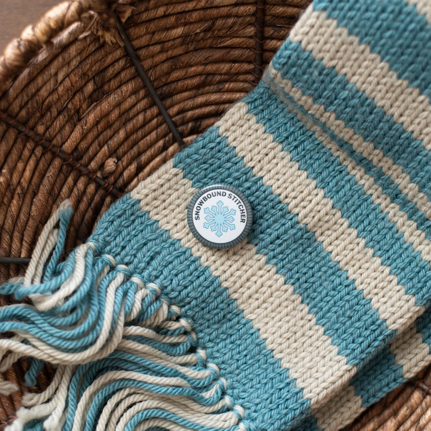 Blue and white striped scarf with a snowbound stitcher merit badge in a woven basket