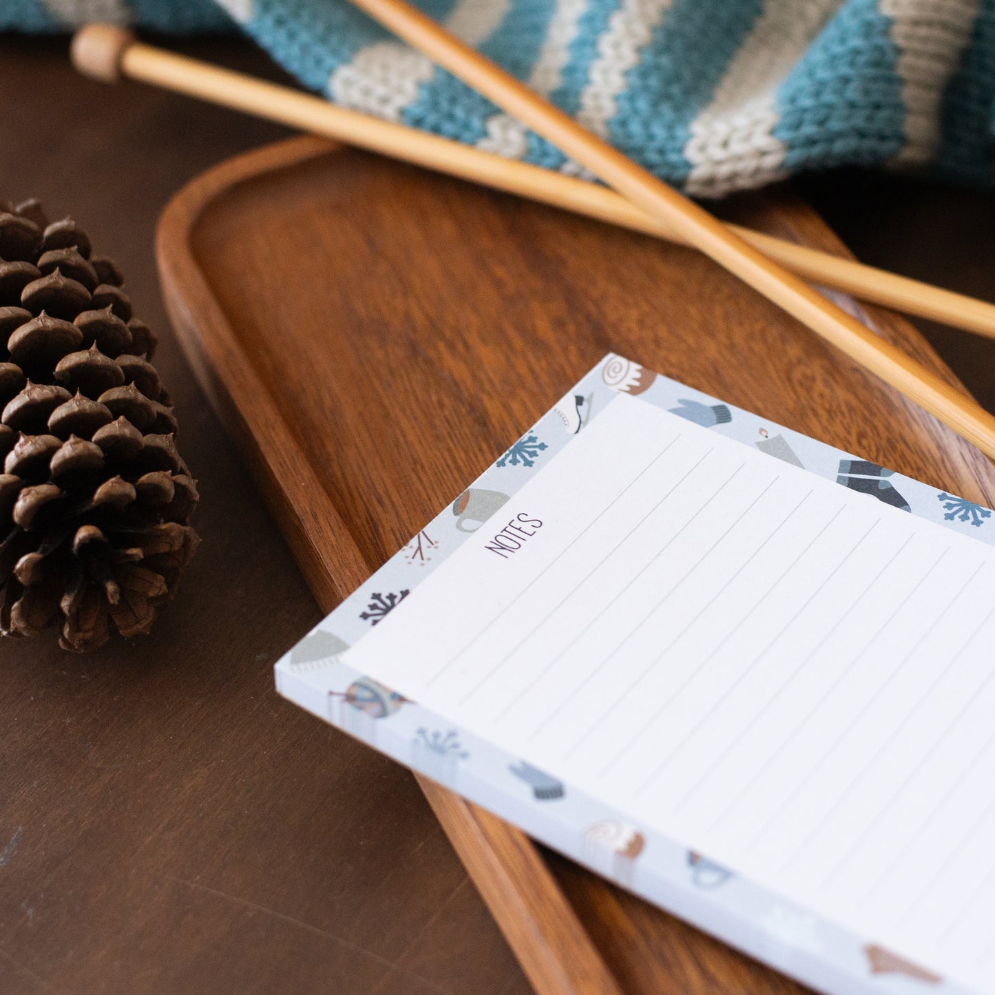 Notepad with decorative cover on a wooden surface with a pine cone and striped scarf.