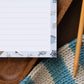 Notepad with a winter-themed design on a wooden surface with a textured scarf.