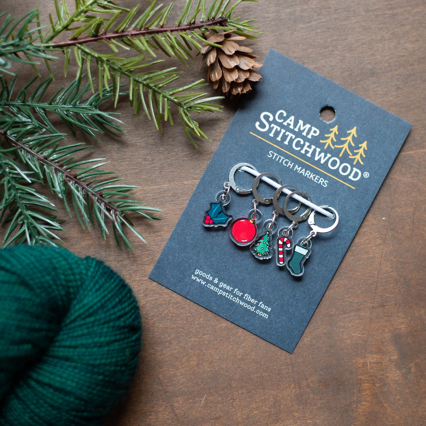 Camp Stitchwood stitch markers on a card with green yarn and pine branch on a wooden surface