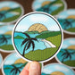 Circular sticker with a tropical design held by a hand, surrounded by more stickers on a brown background.