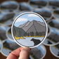 Sticker of a mountain landscape with a bear held over a pile of similar stickers.
