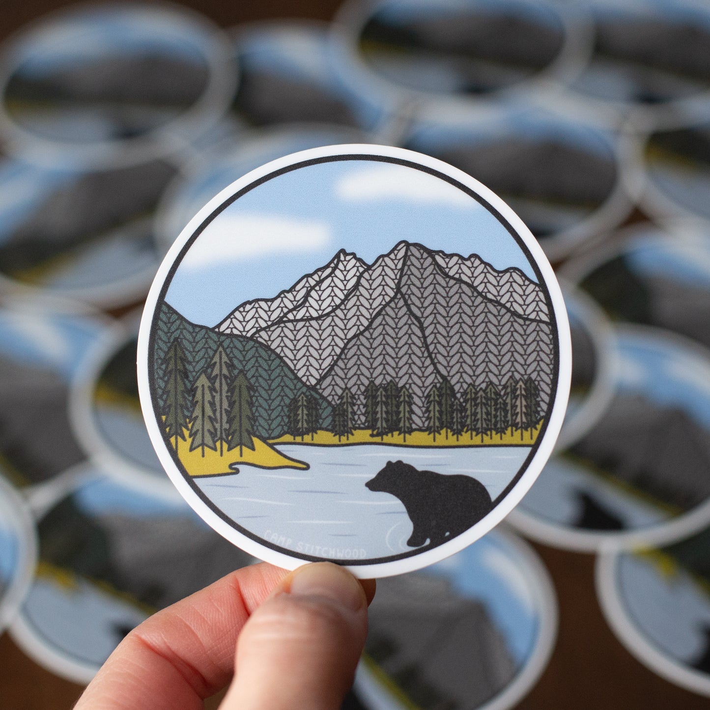 Sticker of a mountain landscape with a bear held over a pile of similar stickers.
