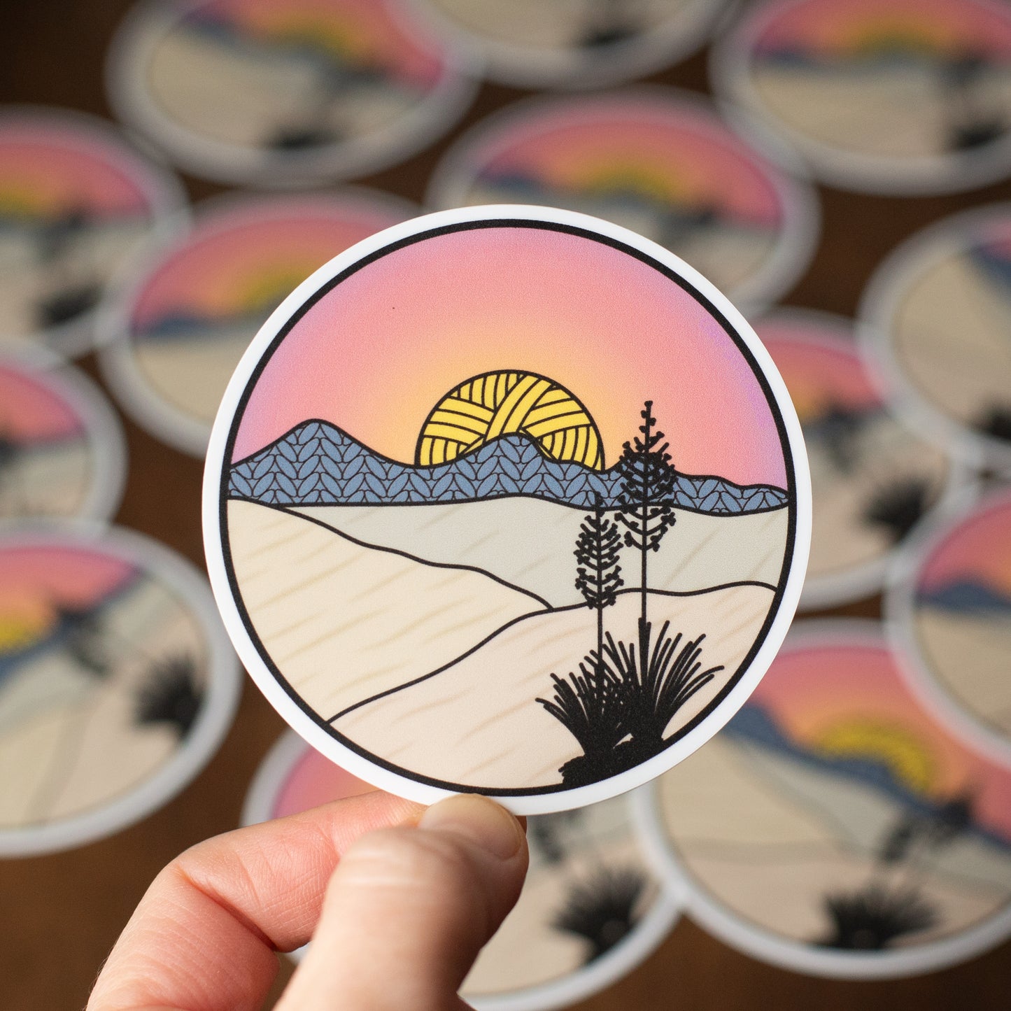 Sticker of a desert landscape with a sunset held by a hand, surrounded by more stickers in the background.