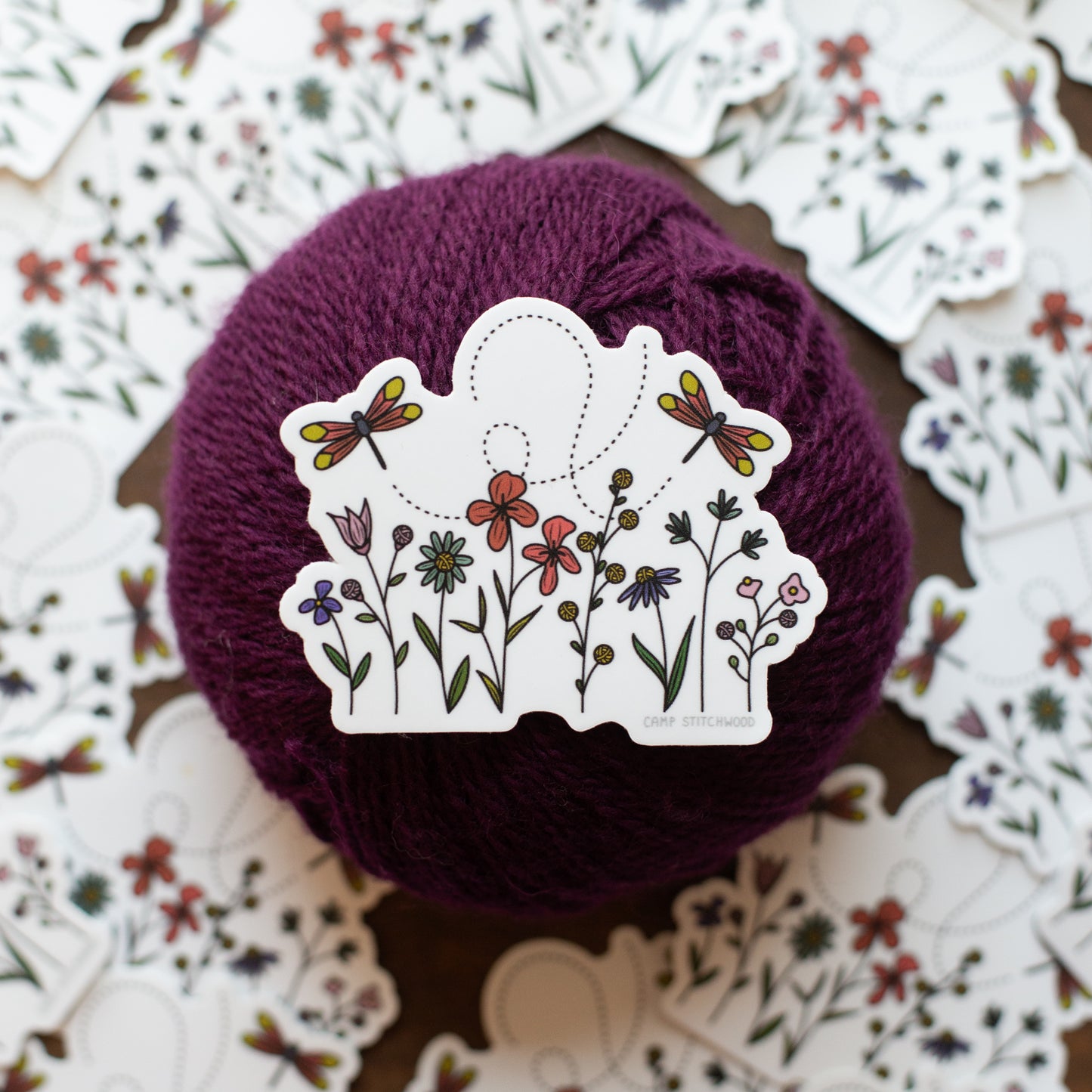 Ball of purple yarn with a decorative sticker featuring flowers and dragonflies on a floral-patterned background.