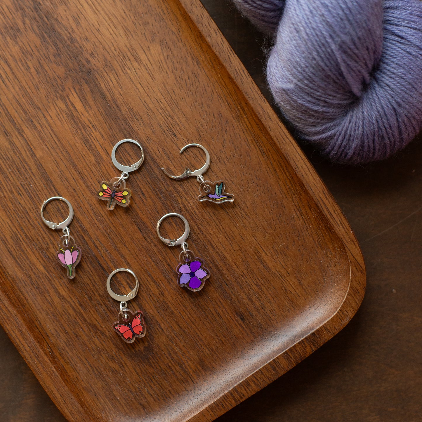 Set of colorful stitch markers charms on a wooden surface with purple yarn.