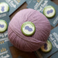 Ball of pink yarn with a 'Social Stitcher' label on a wooden surface with brochures.