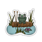 Sticker of a frog sitting on a hank of yarn with grass and water in the background
