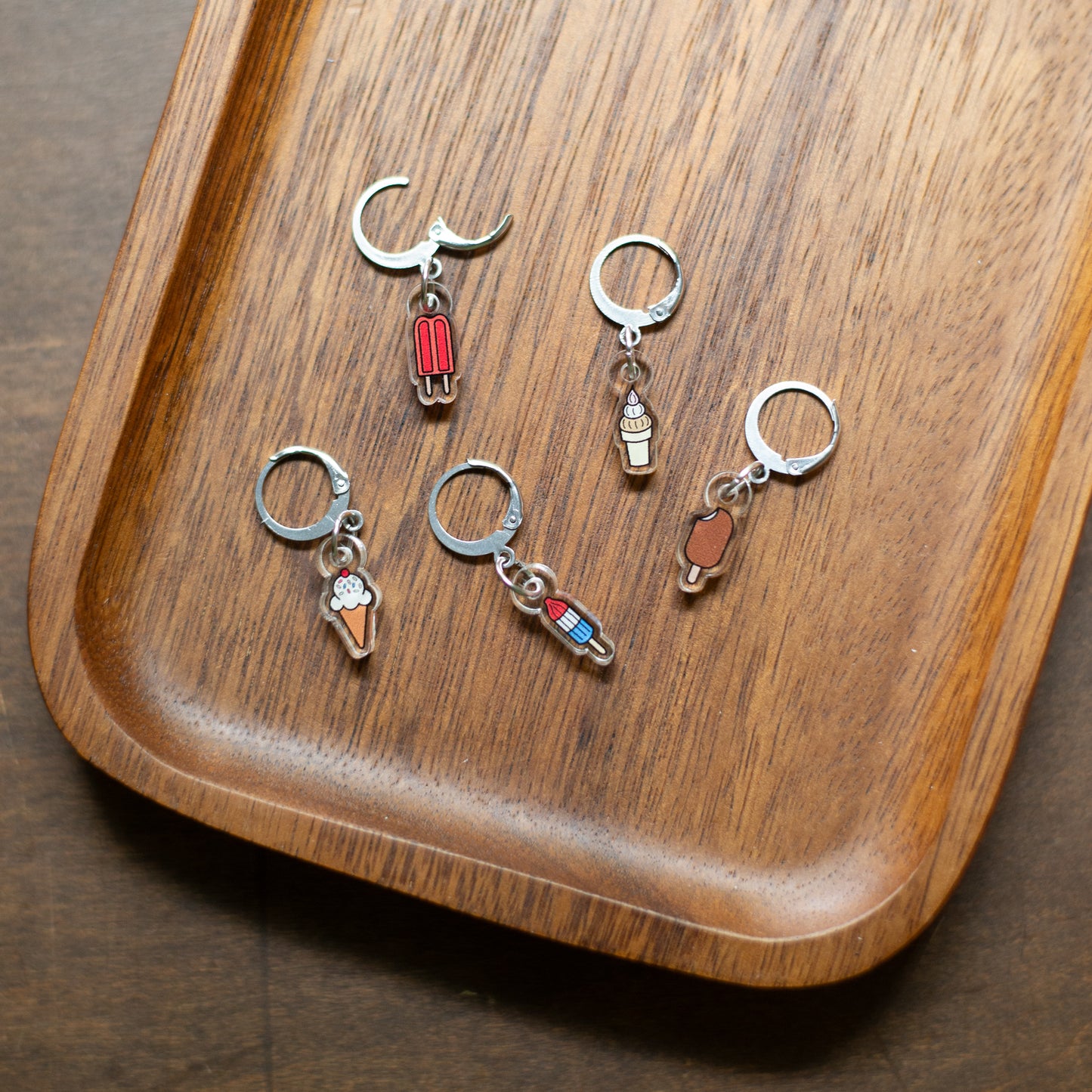 Five stitch markers with small enamel ice cream and popsicle designs, arranged on a curved wooden tray.