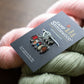 A close-up of a Camp Stitchwood acrylic stitch marker set on its card, resting on a background of pink and sage green yarn skeins.