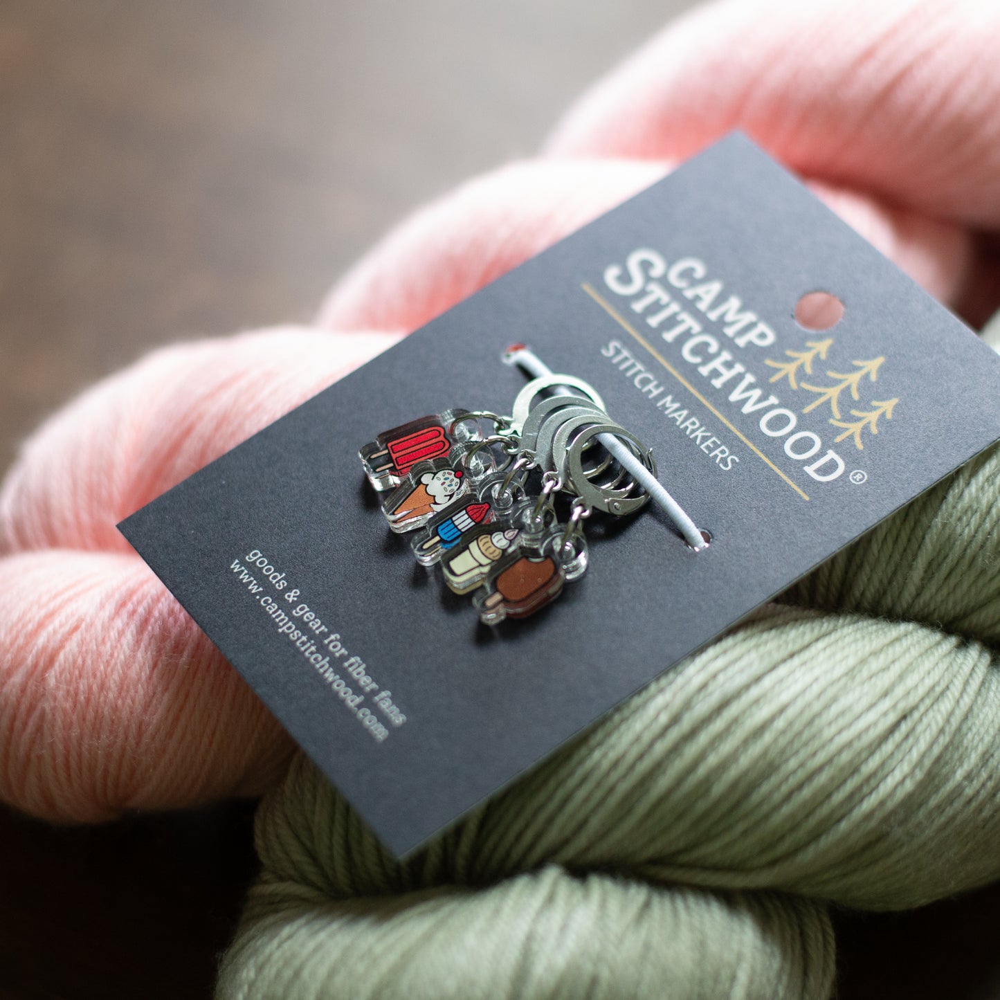A close-up of a Camp Stitchwood acrylic stitch marker set on its card, resting on a background of pink and sage green yarn skeins.