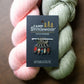 Camp Stitchwood stitch markers on a black display card, styled with pink and sage green yarn skeins on a dark wooden surface.