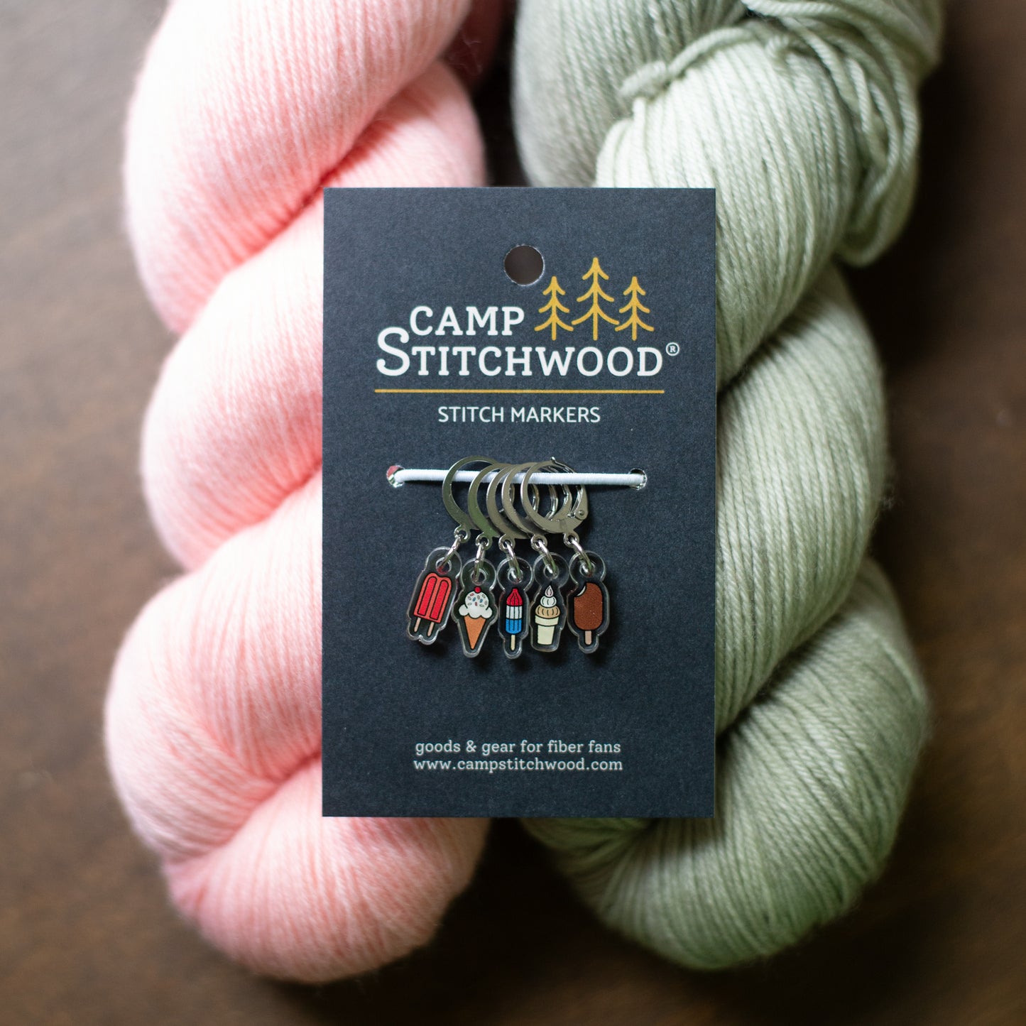 Camp Stitchwood stitch markers on a black display card, styled with pink and sage green yarn skeins on a dark wooden surface.